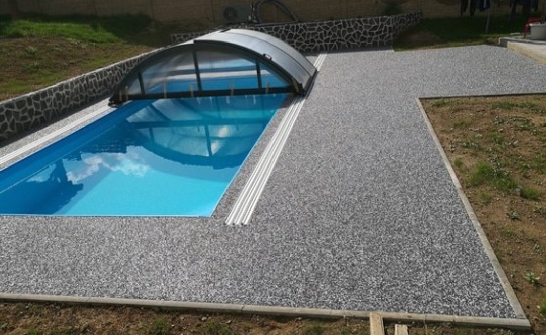 Wykończony teren wokół basenu z szarą nawierzchnią z drobnych kamyczków, częściowo zadaszony basen z błękitną wodą, w tle trawnik i kamienny murek.