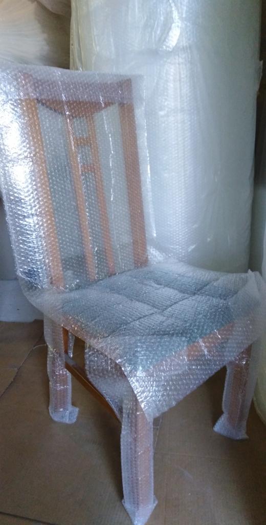 Drewniane krzesło z tapicerowanym siedziskiem zabezpieczone folią bąbelkową, gotowe do transportu, z rolką folii w tle.