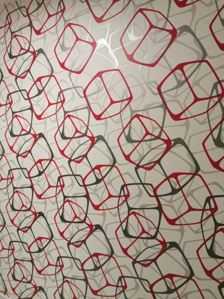 Ściana pokryta tapetą z geometrycznym wzorem czerwonych i szarych obrysów sześcianów na beżowym tle z delikatną teksturą.