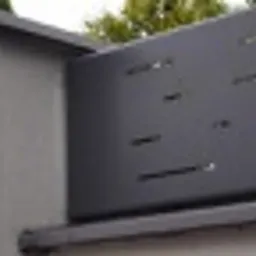 Fragment nowoczesnego ogrodzenia z metalowych paneli w kolorze grafitowym, zamontowanego na betonowym słupku z szarym daszkiem. Panele z poziomymi, prostokątnymi wycięciami. Widok z bliska, lekko...