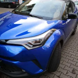 Niebieski samochód typu crossover, Toyota C-HR Hybrid, zaparkowany na brukowanej ulicy przed budynkiem z szyldem sklepu.