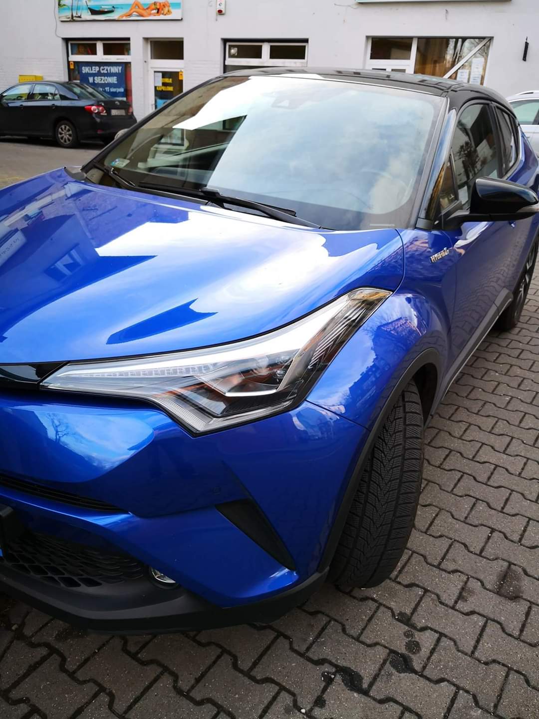 Niebieski samochód typu crossover, Toyota C-HR Hybrid, zaparkowany na brukowanej ulicy przed budynkiem z szyldem sklepu.