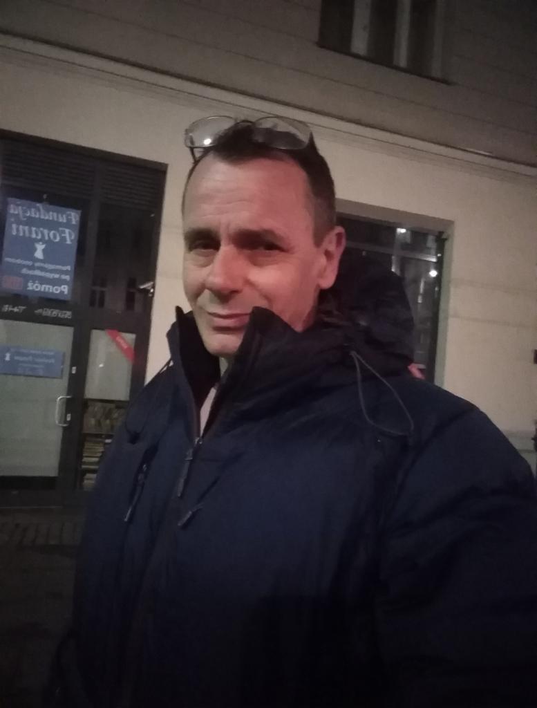 Portret mężczyzny w kurtce na tle budynku z oknami i napisem, okulary na głowie.