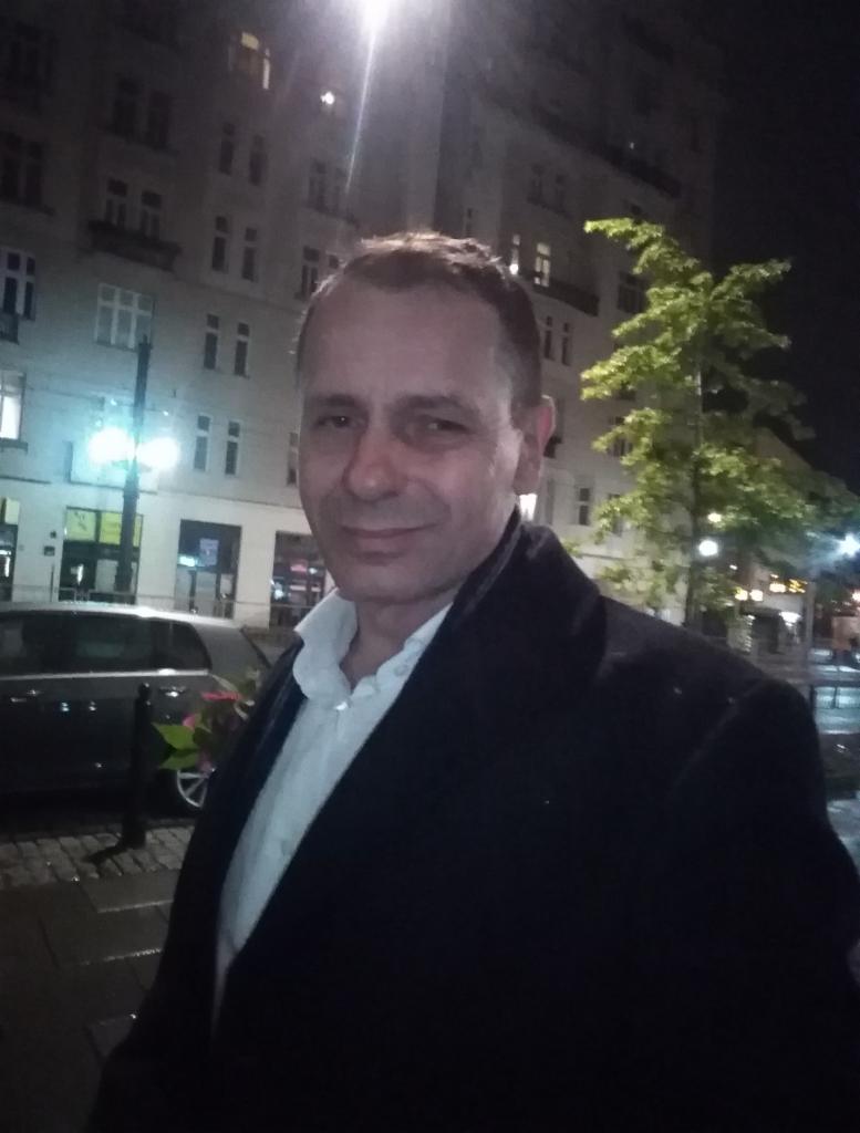 Portret mężczyzny w ciemnym płaszczu i białej koszuli na tle miejskiej ulicy nocą, z widoczną kamienicą i oświetleniem ulicznym.