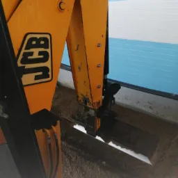 Żółta koparka JCB w trakcie wykonywania wykopu w Kamieńcu, widoczne świeżo wykopane podłoże i fragment hali w tle.