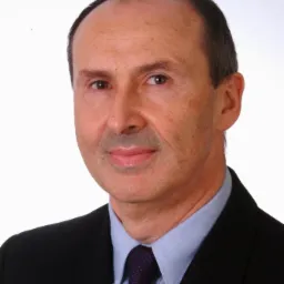 Portret mężczyzny w garniturze i krawacie z subtelnym uśmiechem na jasnym tle.