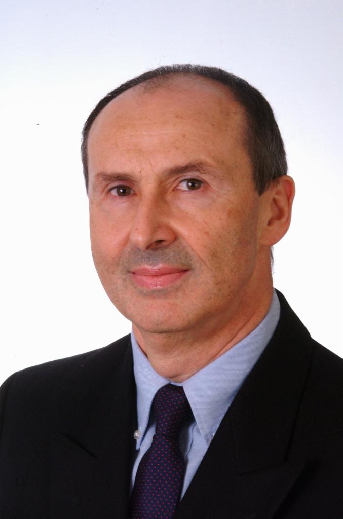 Portret mężczyzny w garniturze i krawacie z subtelnym uśmiechem na jasnym tle.