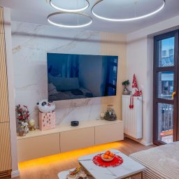 Kasperek Jan - Nowoczesny salon z podświetlaną szafką RTV, marmurową ścianą za telewizorem, okrągłymi lampami wiszącymi i dekoracjami świątecznymi na grzejniku.