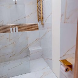 Kasperek Jan - Nowoczesna łazienka z marmurowymi płytkami w odcieniach bieli i złota, złoty grzejnik drabinkowy, szklana kabina prysznicowa, półka z trzema białymi butelkami kosmetyków i uchwyt na papier...