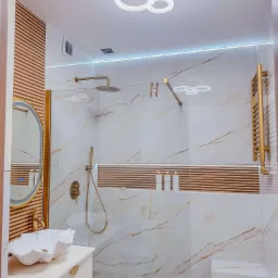 Nowoczesna łazienka z marmurowymi płytkami, złotą armaturą, umywalką w kształcie muszli i oświetleniem LED, akcentowana drewnianymi panelami i geometryczną lampą sufitową.