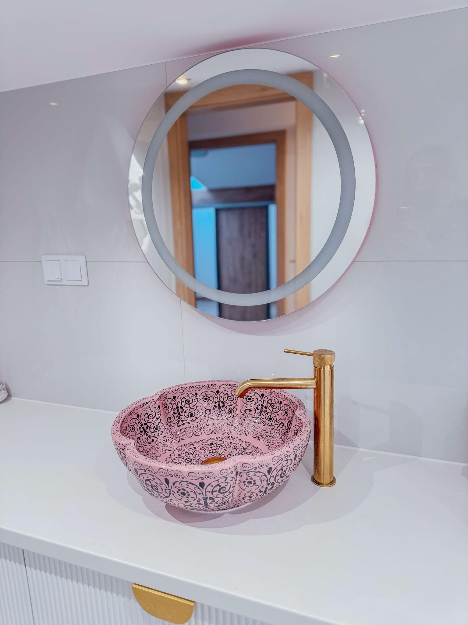 Umywalka ceramiczna w kolorze pudrowego różu z czarnym ornamentem, złota bateria umywalkowa, okrągłe lustro z podświetleniem LED w łazience wykończonej białymi płytkami.