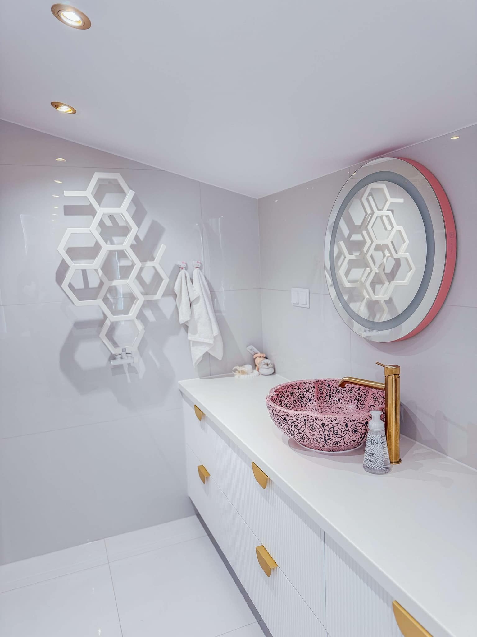 Nowoczesna łazienka z umywalką w kolorze pudrowego różu z czarnym wzorem, złotą baterią, lustrem z podświetleniem LED w kształcie plastra miodu i białymi meblami z frezowanymi frontami.