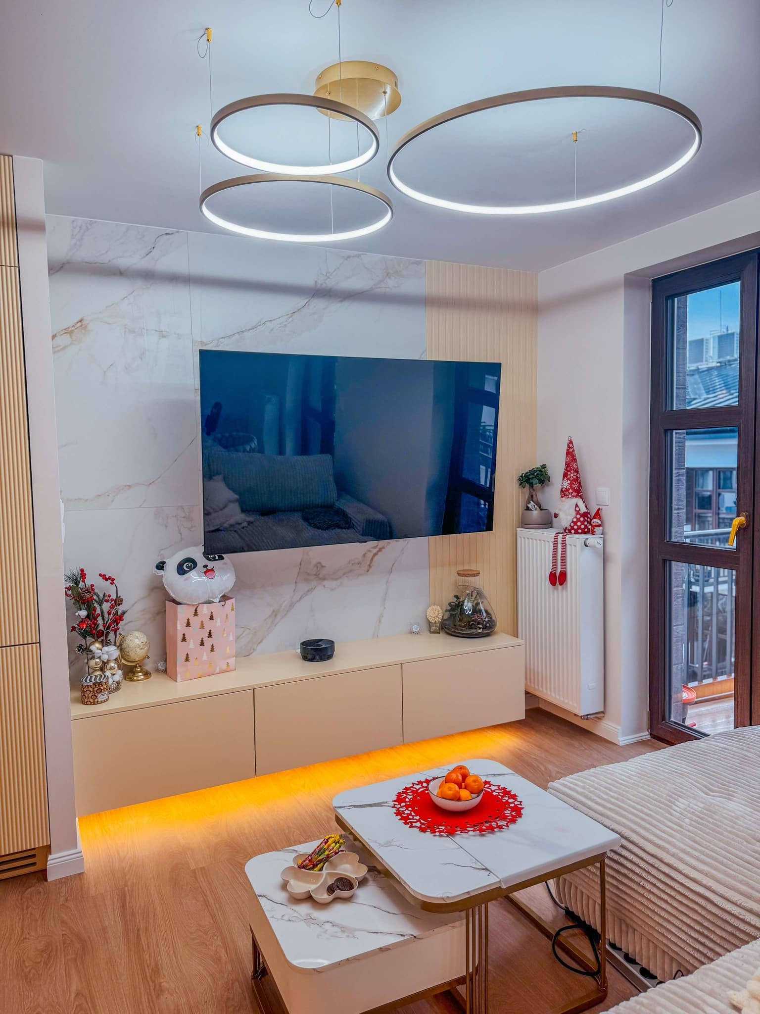 Nowoczesny salon z podświetlaną szafką RTV, marmurową ścianą za telewizorem, okrągłymi lampami wiszącymi i dekoracjami świątecznymi na grzejniku.