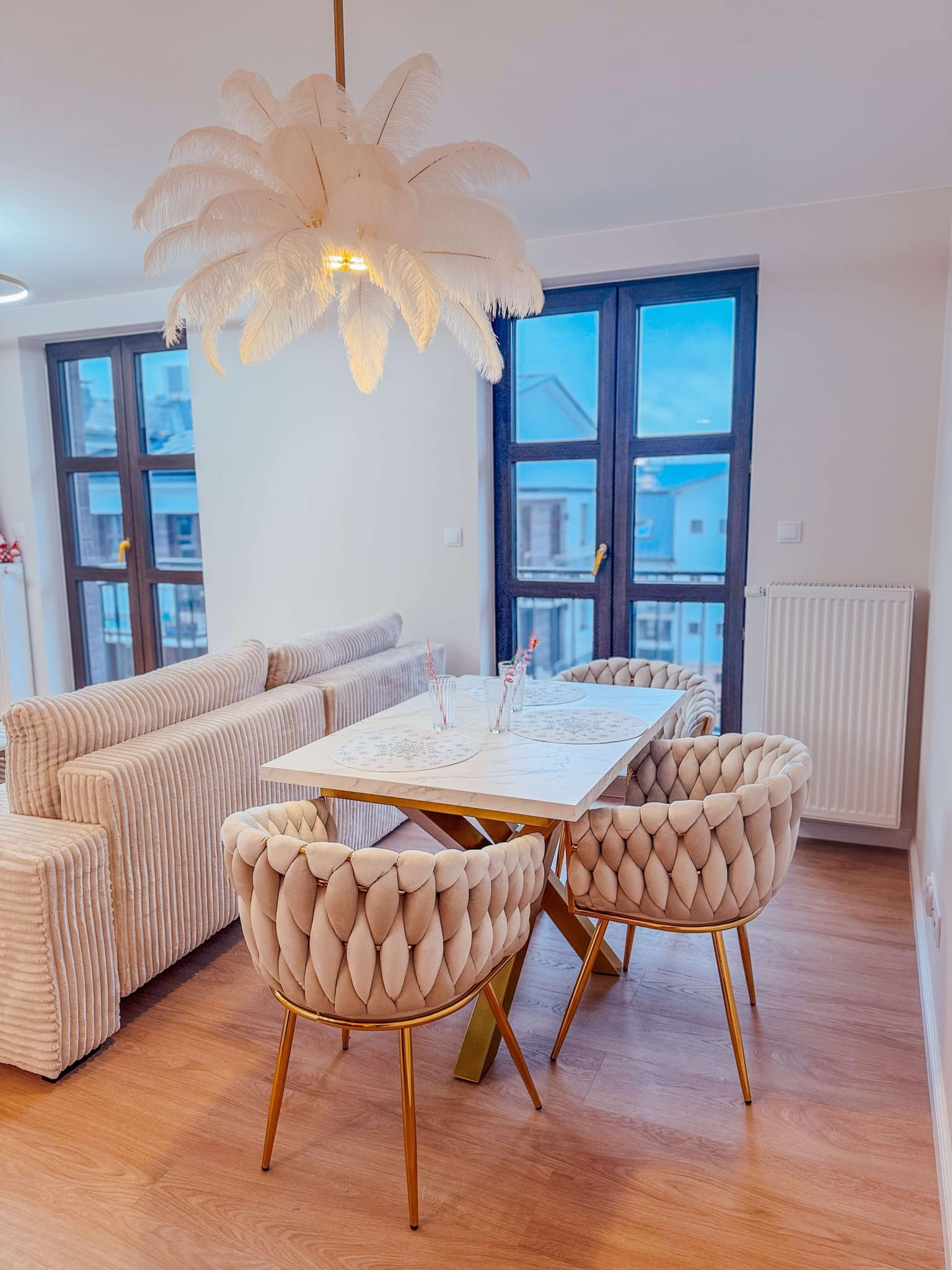 Jasne wnętrze jadalni z beżową sofą, białym stołem na złotych nogach, beżowymi krzesłami z plecionym oparciem na złotych nogach i lampą w kształcie pióropusza.