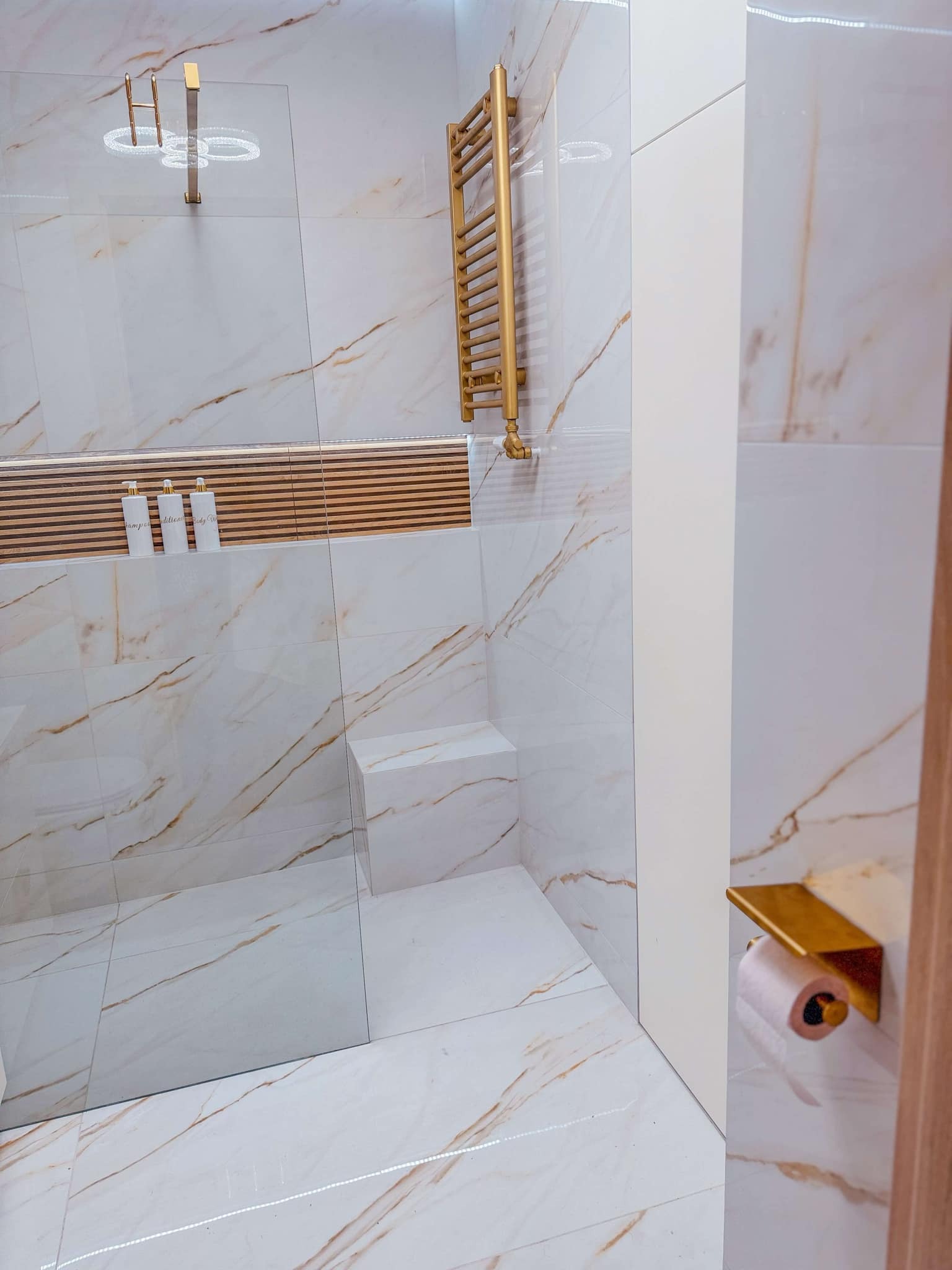 Nowoczesna łazienka z marmurowymi płytkami w odcieniach bieli i złota, złoty grzejnik drabinkowy, szklana kabina prysznicowa, półka z trzema białymi butelkami kosmetyków i uchwyt na papier...