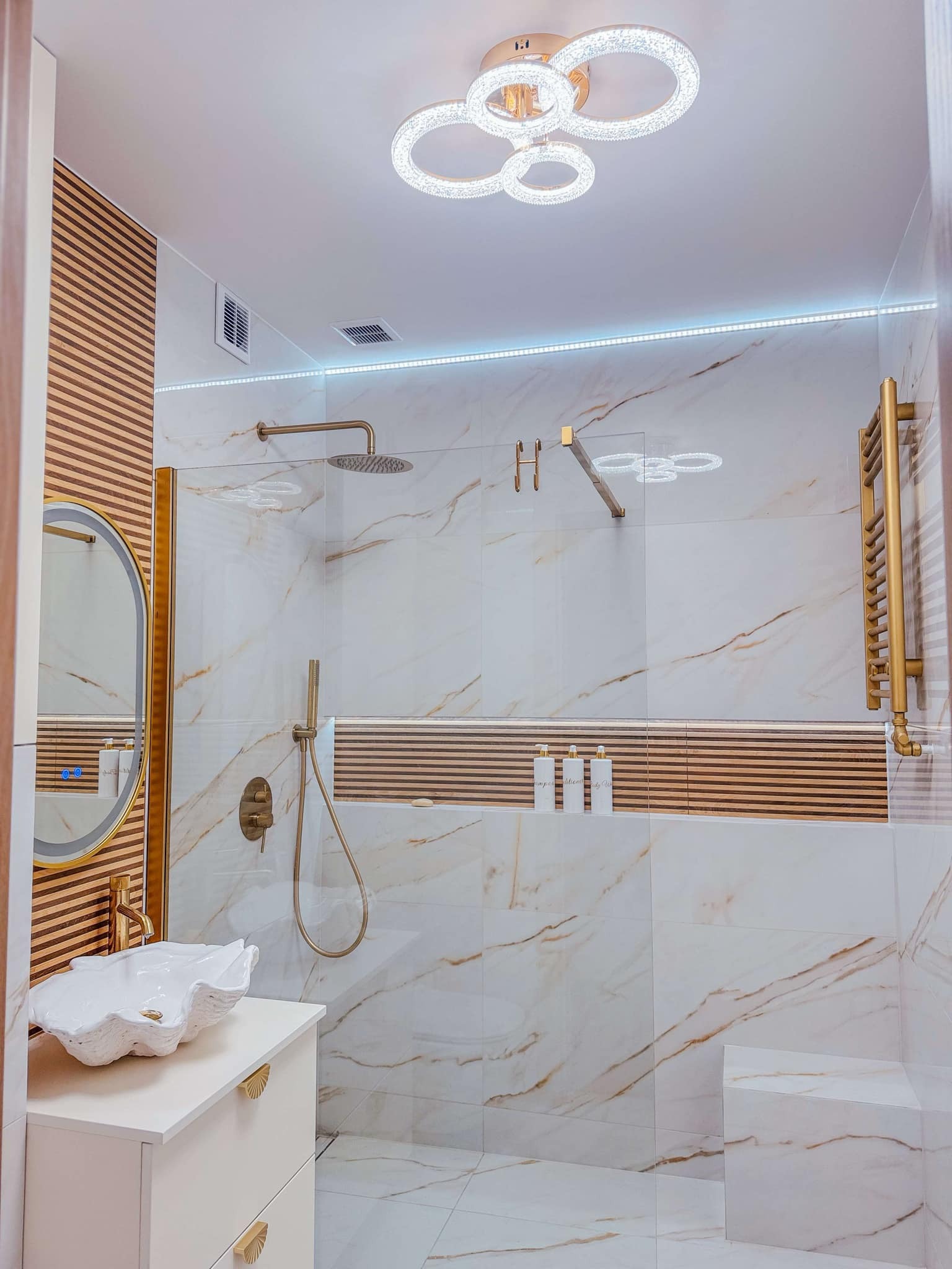 Nowoczesna łazienka z marmurowymi płytkami, złotą armaturą, umywalką w kształcie muszli i oświetleniem LED, akcentowana drewnianymi panelami i geometryczną lampą sufitową.