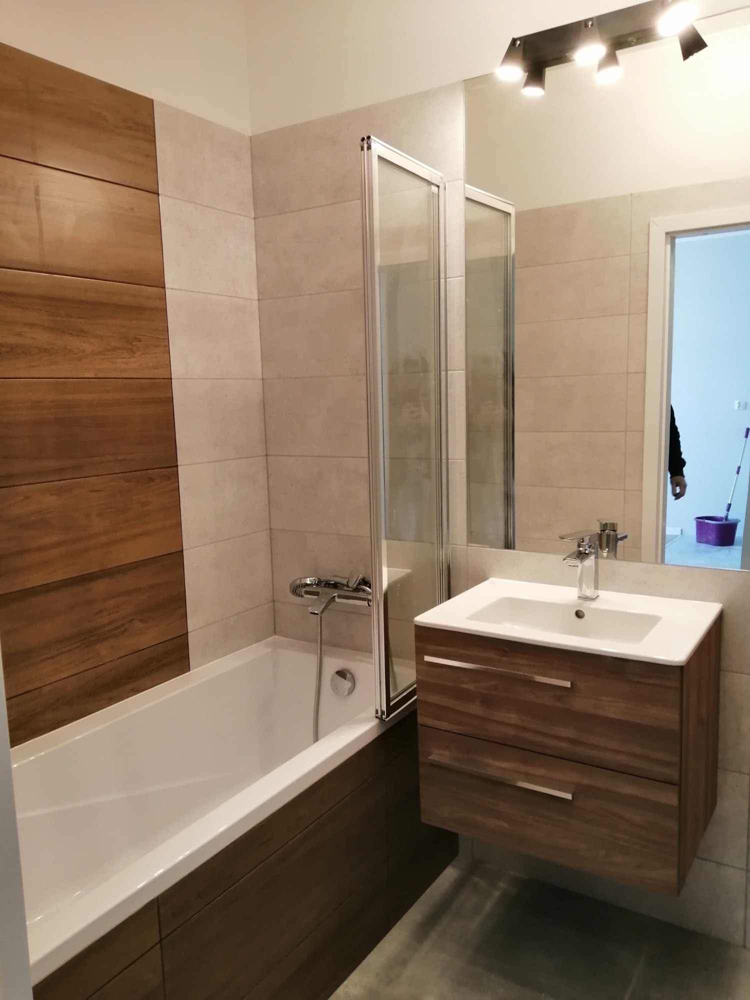 Nowoczesna łazienka z wanną obudowaną drewnopodobnymi panelami, szklanym parawanem nawannowym i szafką pod umywalką z dwoma szufladami, widok na fragment osoby sprzątającej w lustrze.