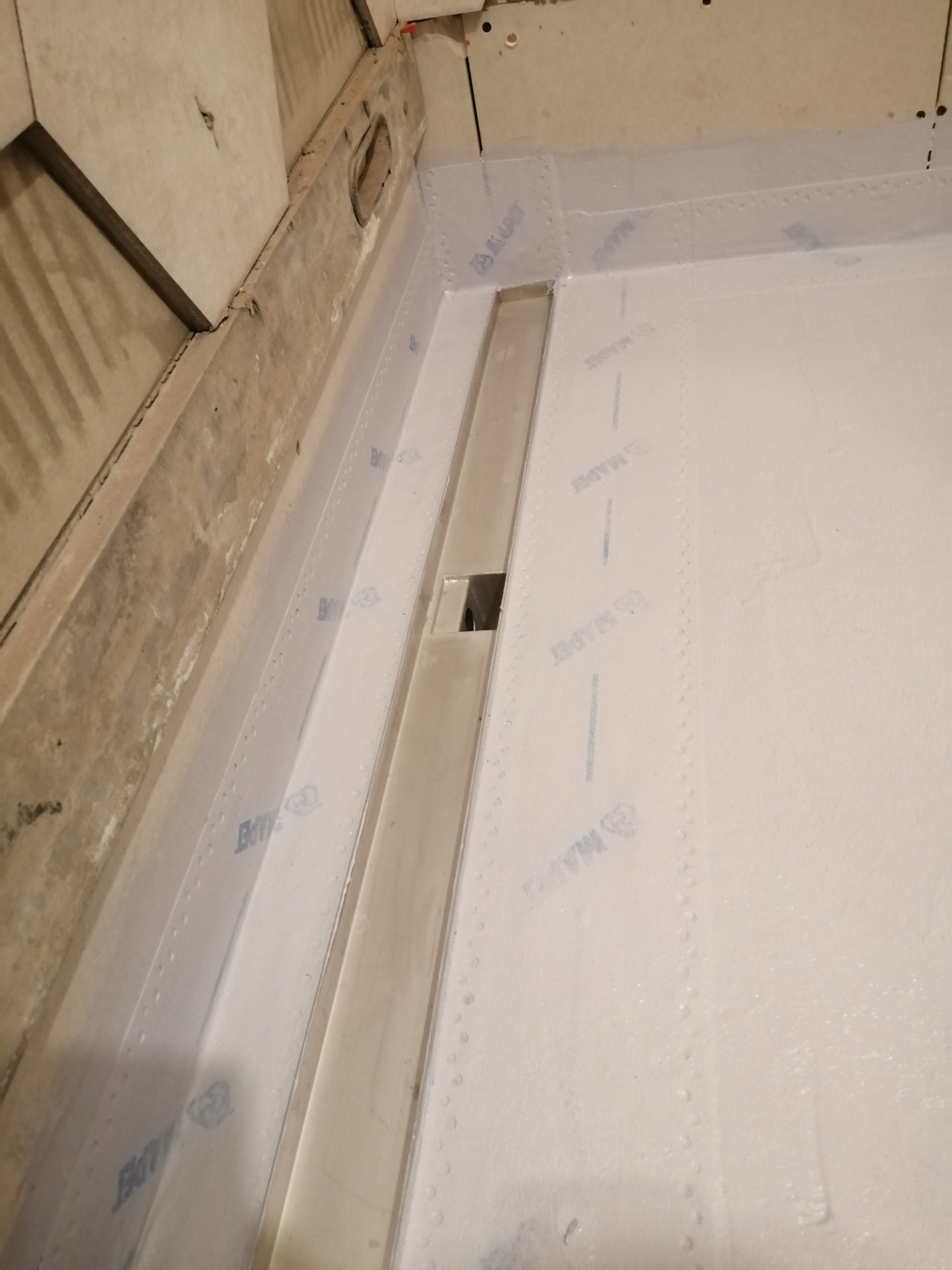 Przygotowanie podłoża pod prysznic z odpływem liniowym, zabezpieczone folią w płynie z widocznym logo producenta, surowy beton na ścianie.