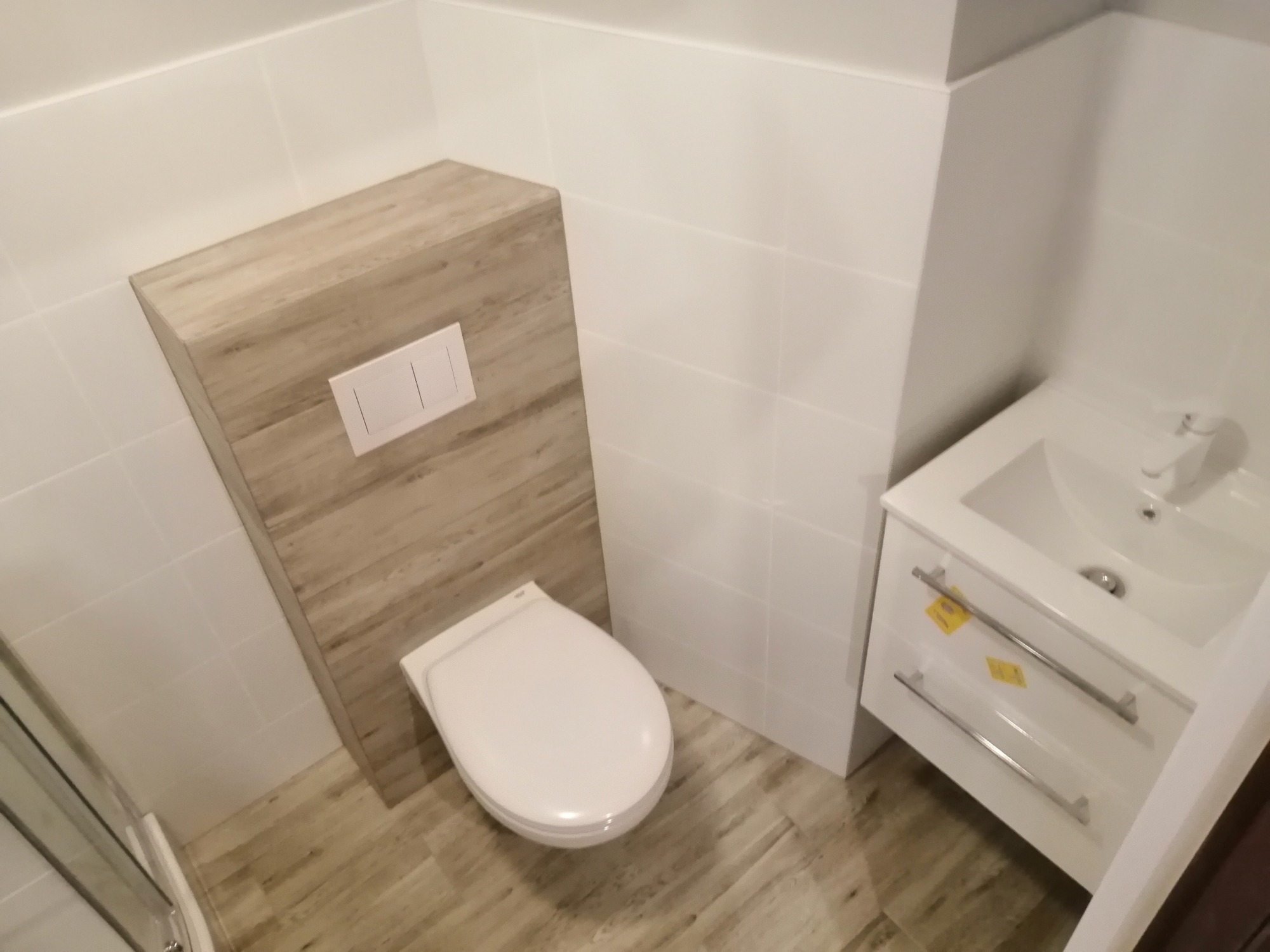 Nowoczesna łazienka po remoncie: podwieszana toaleta z drewnianą obudową stelaża i biała umywalka z szafką, jasne płytki na ścianach i podłodze imitujące drewno.