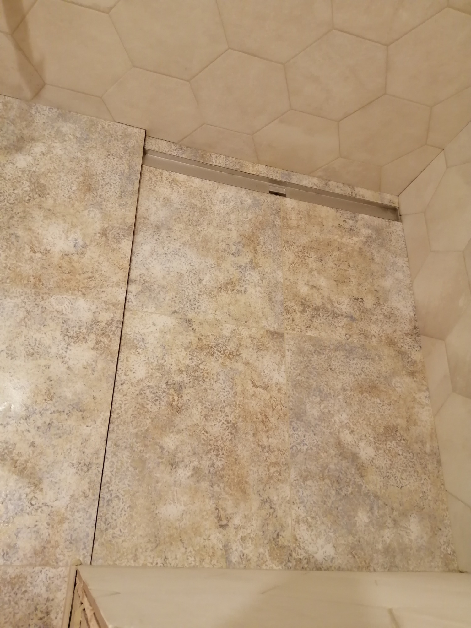 Fragment ściany w łazience wyłożonej beżowymi płytkami z delikatnym wzorem, widoczny odpływ liniowy i heksagonalne płytki na suficie.