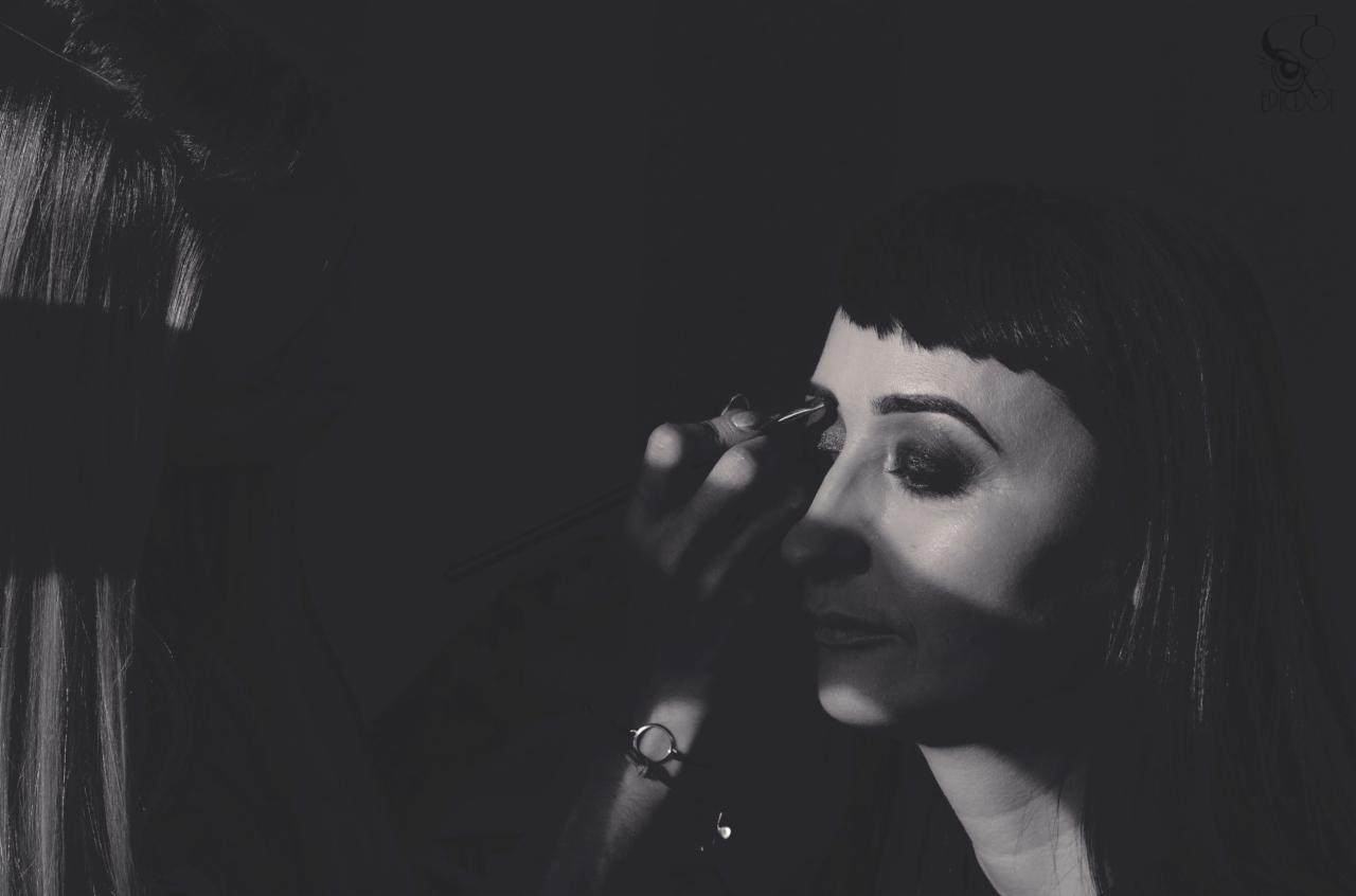 Czarno-białe zdjęcie profilu kobiety podczas regulacji brwi pęsetą, z widocznym makijażem oczu i zegarkiem na nadgarstku osoby wykonującej zabieg.