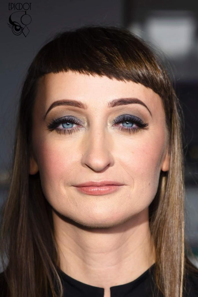 Portret kobiety z subtelnym makijażem oczu w odcieniach szarości i brązu, podkreślonymi sztucznymi rzęsami, oraz delikatnym makijażem ust, z charakterystyczną krótką grzywką i jasnymi tęczówkami.