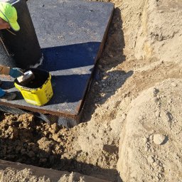 NJAA JANUSZ BARANOWSKI SPÓŁKA Z OGRANICZONĄ ODPOWIEDZIALNOŚCIĄ - Montaż betonowego zbiornika: pracownik z żółtym wiaderkiem i pędzlem nanosi uszczelnienie na zewnętrzną powierzchnię szamba, widoczny wykop w ziemi z zabezpieczeniem hydroizolacyjnym.