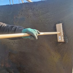 NJAA JANUSZ BARANOWSKI SPÓŁKA Z OGRANICZONĄ ODPOWIEDZIALNOŚCIĄ - Hydroizolacja zewnętrznej ściany szamba betonowego, nałożona pędzlem na długim trzonku, widoczne mocowania do transportu dźwigiem.