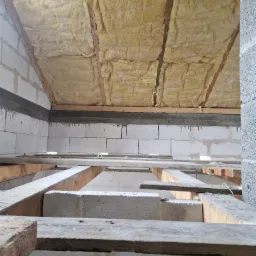 Wnętrze budynku w trakcie budowy, widoczne bloczki betonowe, drewniane belki stropowe oraz izolacja z wełny mineralnej pod dachem.