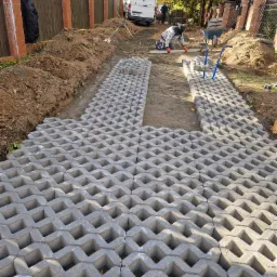 Układanie ażurowych płyt betonowych na przygotowanym podłożu, widoczny pracownik w pomarańczowych rękawicach poziomujący powierzchnię, w tle ziemia, taczka i zaparkowany biały samochód dostawczy.