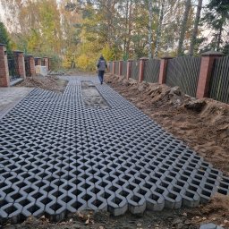 NJAA JANUSZ BARANOWSKI SPÓŁKA Z OGRANICZONĄ ODPOWIEDZIALNOŚCIĄ - Układanie szarej ażurowej nawierzchni typu kratka na podjeździe w trakcie realizacji, widoczny fragment ogrodzenia z cegły i drewniane sztachety w tle, jesienne drzewa bez liści.