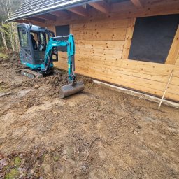 NJAA JANUSZ BARANOWSKI SPÓŁKA Z OGRANICZONĄ ODPOWIEDZIALNOŚCIĄ - Niebieska mini koparka Sunward 20P wykonuje wykop przy drewnianym budynku z zabezpieczonymi oknami, widoczne ślady opon i świeżo naruszona ziemia.