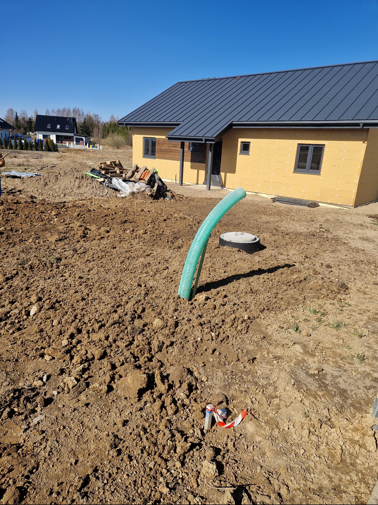 Teren przygotowany pod budowę domu, widoczny wykop z wystającą zieloną rurą, brązowa ziemia, fragment budynku z szarą dachówką i żółtą elewacją, niebieskie niebo.