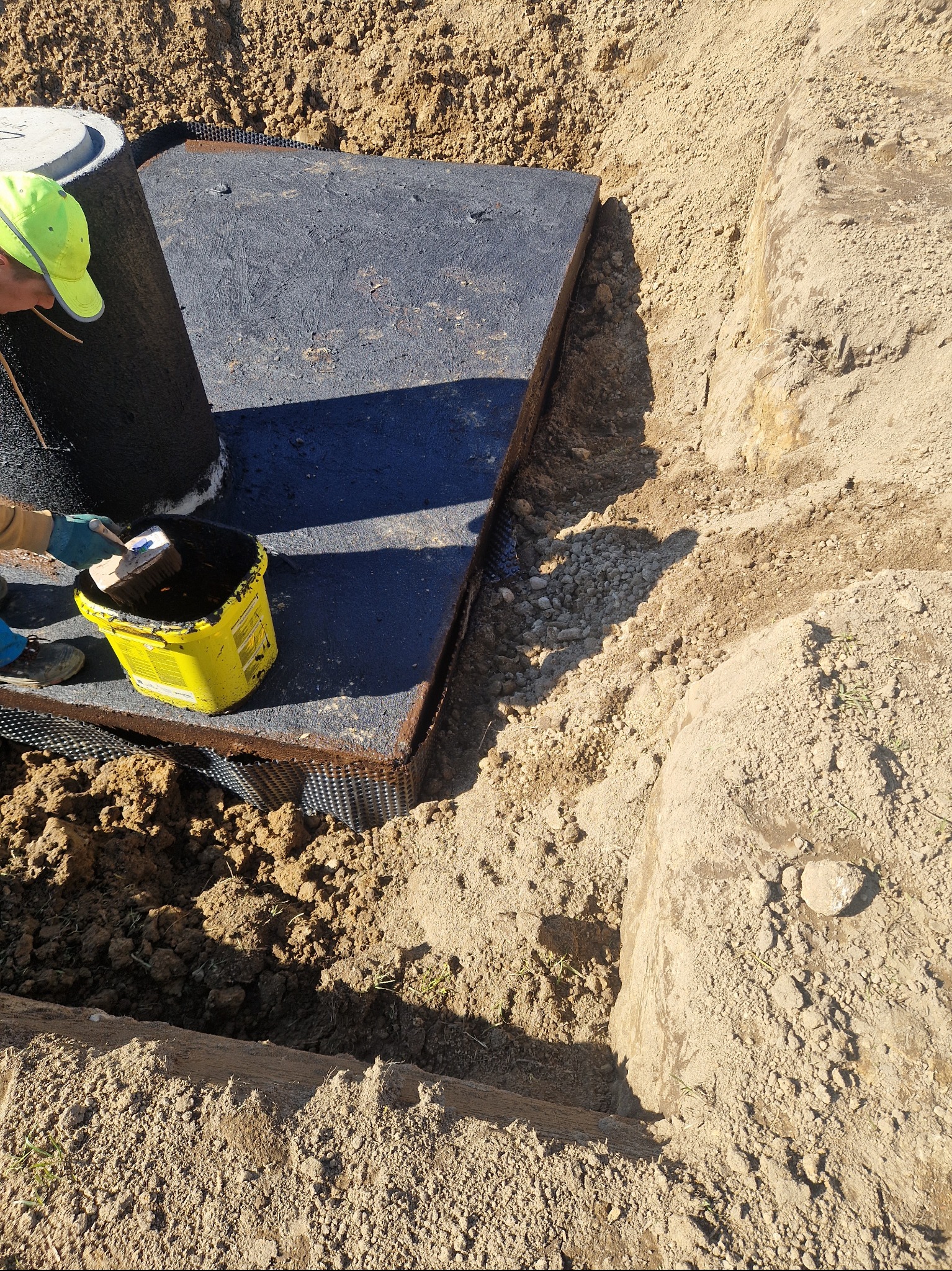 Montaż betonowego zbiornika: pracownik z żółtym wiaderkiem i pędzlem nanosi uszczelnienie na zewnętrzną powierzchnię szamba, widoczny wykop w ziemi z zabezpieczeniem hydroizolacyjnym.