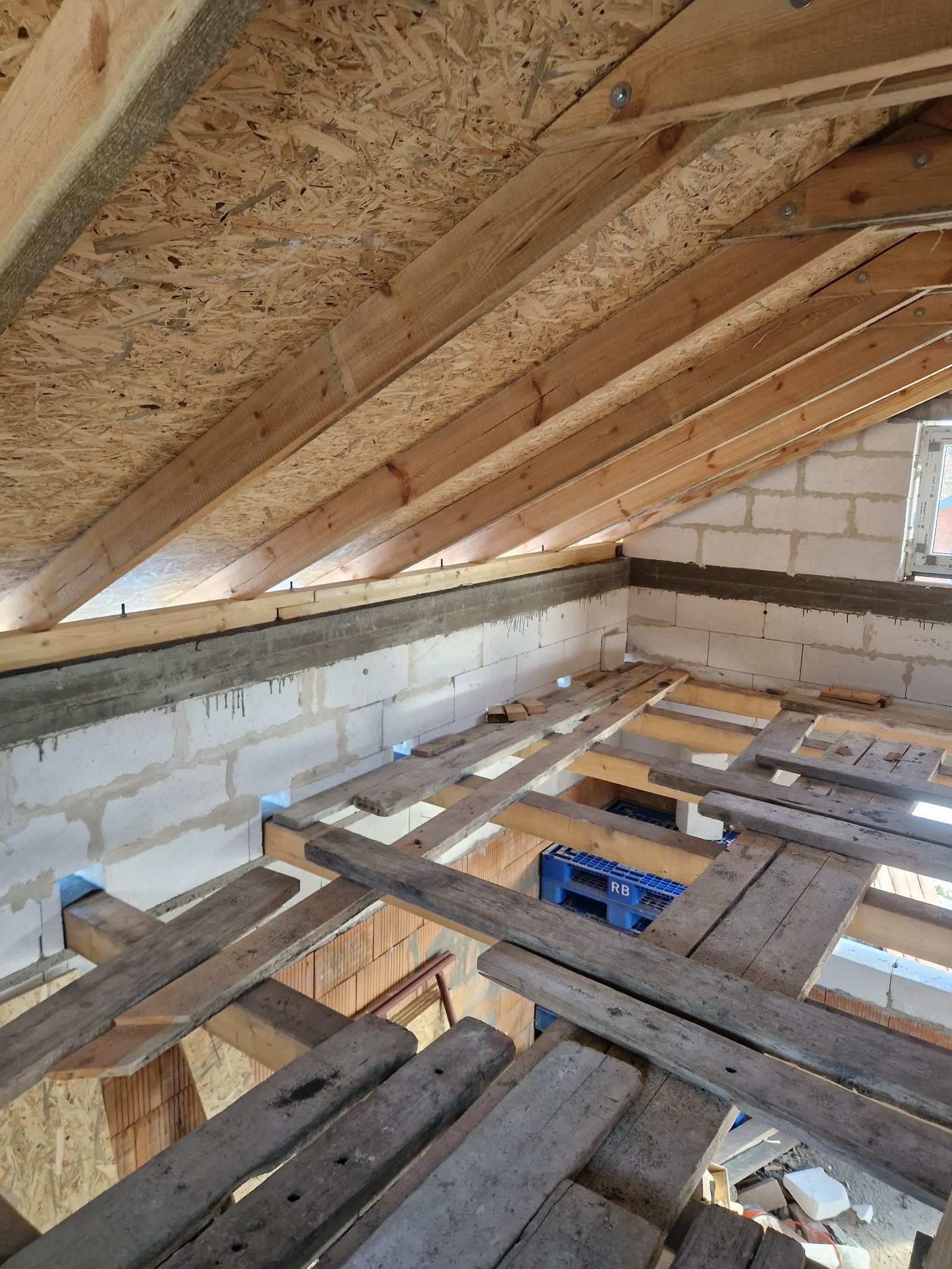 Surowe wnętrze poddasza w trakcie budowy: widoczne białe bloczki, drewniana konstrukcja dachu z płyt OSB, prowizoryczna podłoga z desek, niebieskie plastikowe skrzynki w tle.