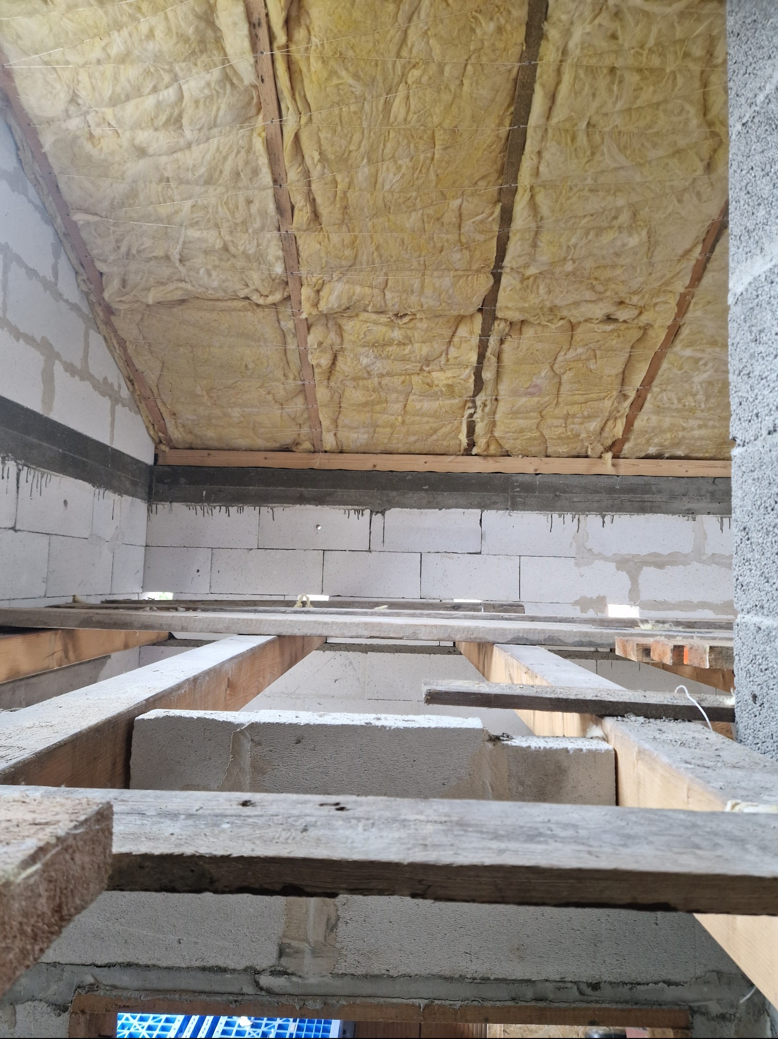 Wnętrze budynku w trakcie budowy, widoczne bloczki betonowe, drewniane belki stropowe oraz izolacja z wełny mineralnej pod dachem.