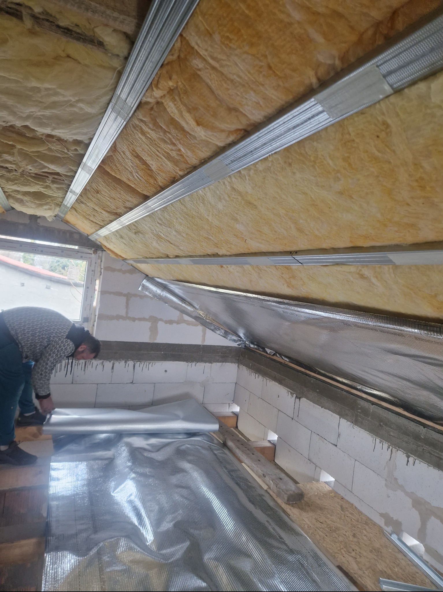 Pracownik montuje folię termoizolacyjną na poddaszu, widoczne krokwie z wełną mineralną i metalowe profile, okno w tle.