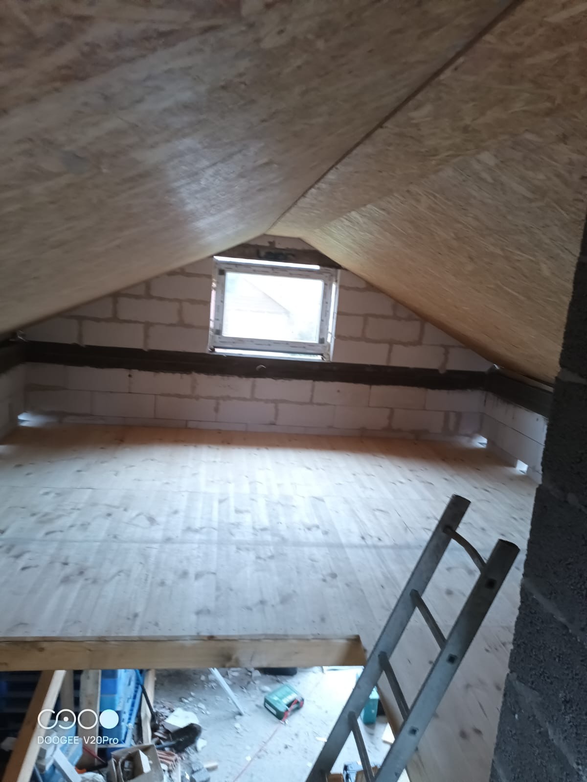 Surowe poddasze z drewnianą podłogą, oknem w szczycie i widoczną drabiną, ściany z bloczków betonowych, widok w trakcie prac wykończeniowych.