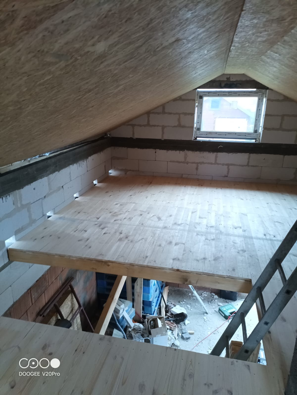 Surowe drewniane deski podłogowe na poddaszu z oknem, widoczne bloczki betonowe i fragment drabiny, w tle gruz i materiały budowlane.
