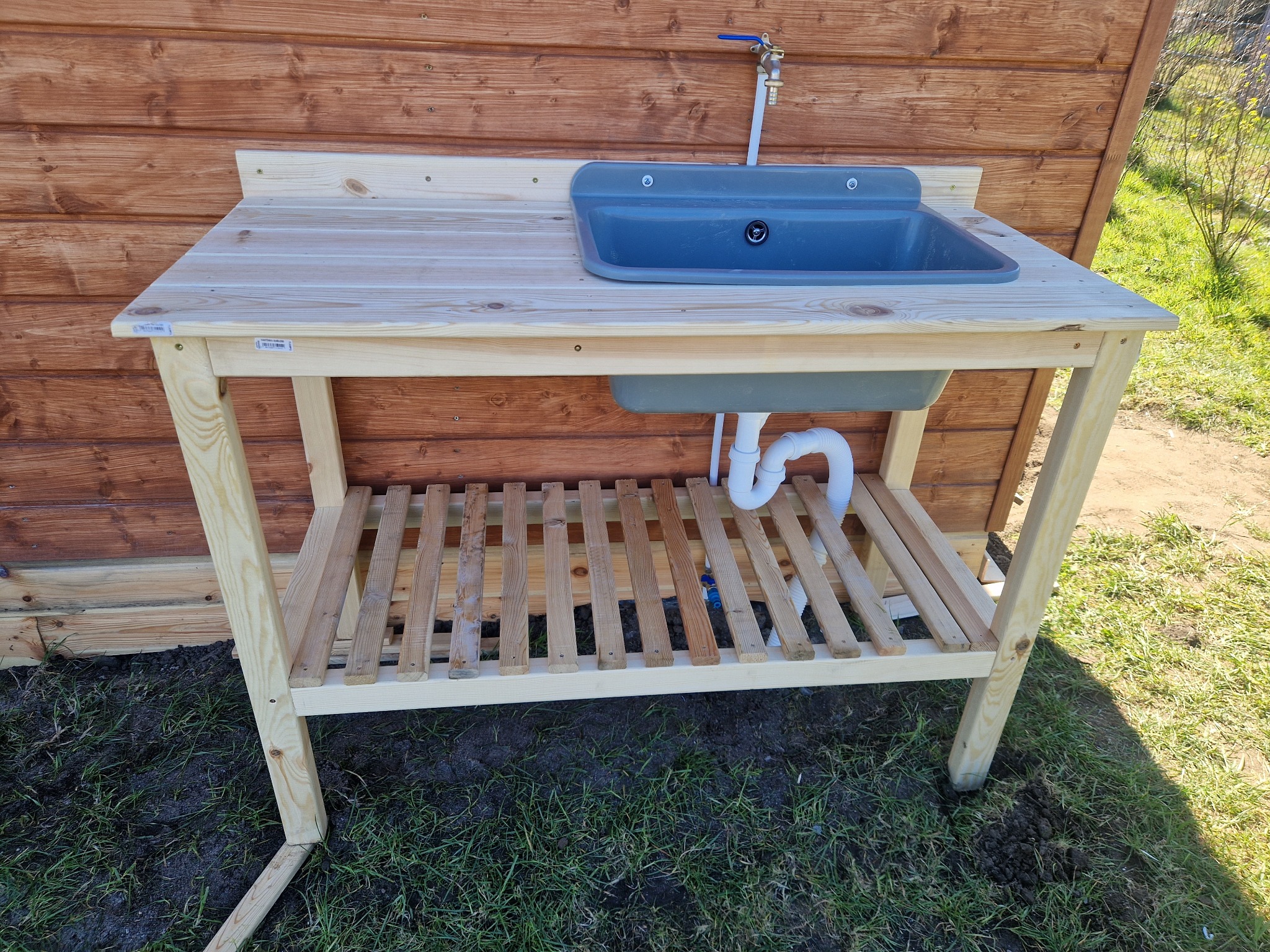 Drewniany stół z szarą umywalką i kranem na zewnątrz, z półką pod spodem, ustawiony na trawie przed drewnianą ścianą.