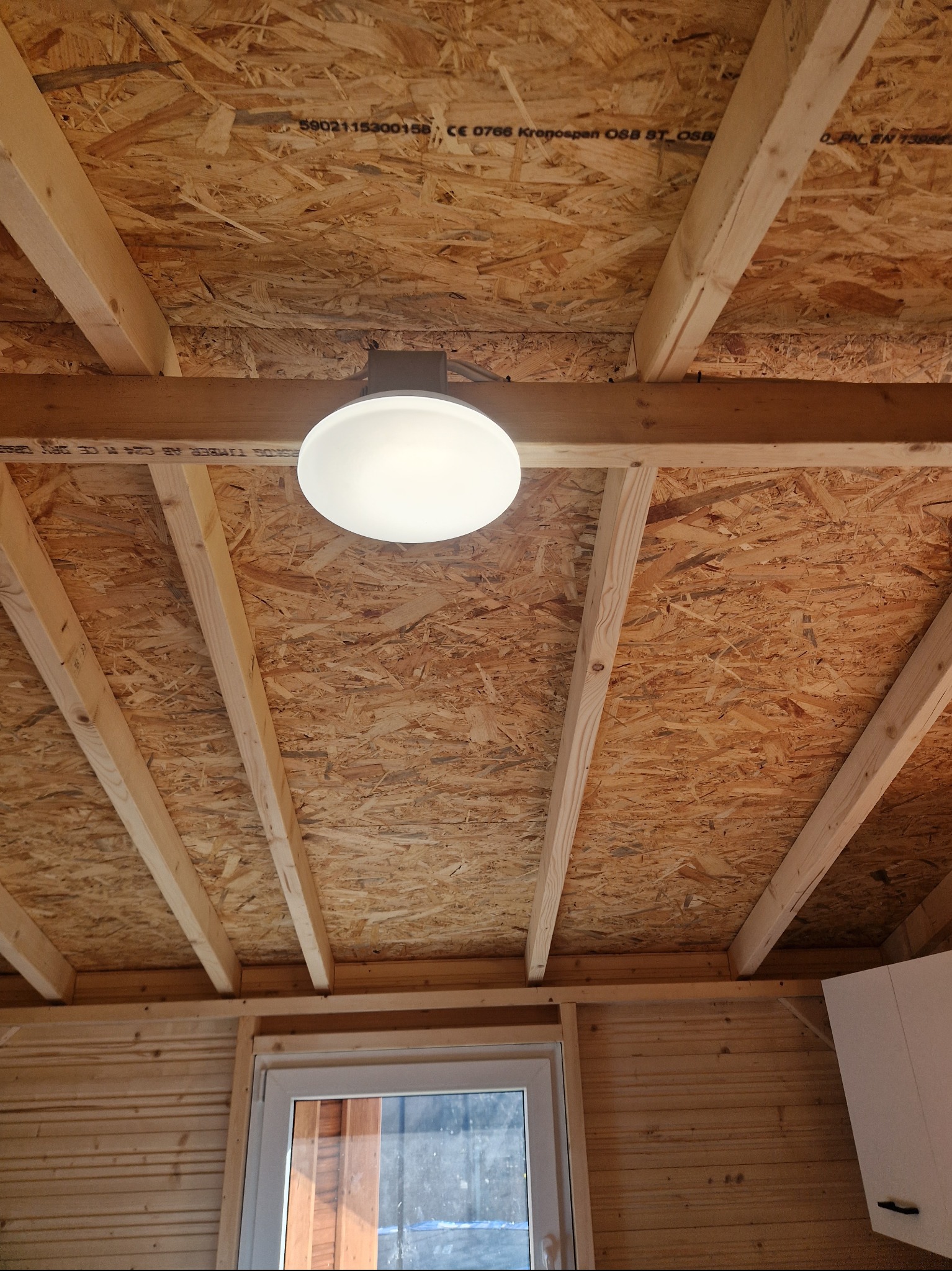 Wnętrze szopy z widocznym drewnianym stropem wykończonym płytą OSB, oświetlone okrągłą lampą sufitową, z oknem w tle.