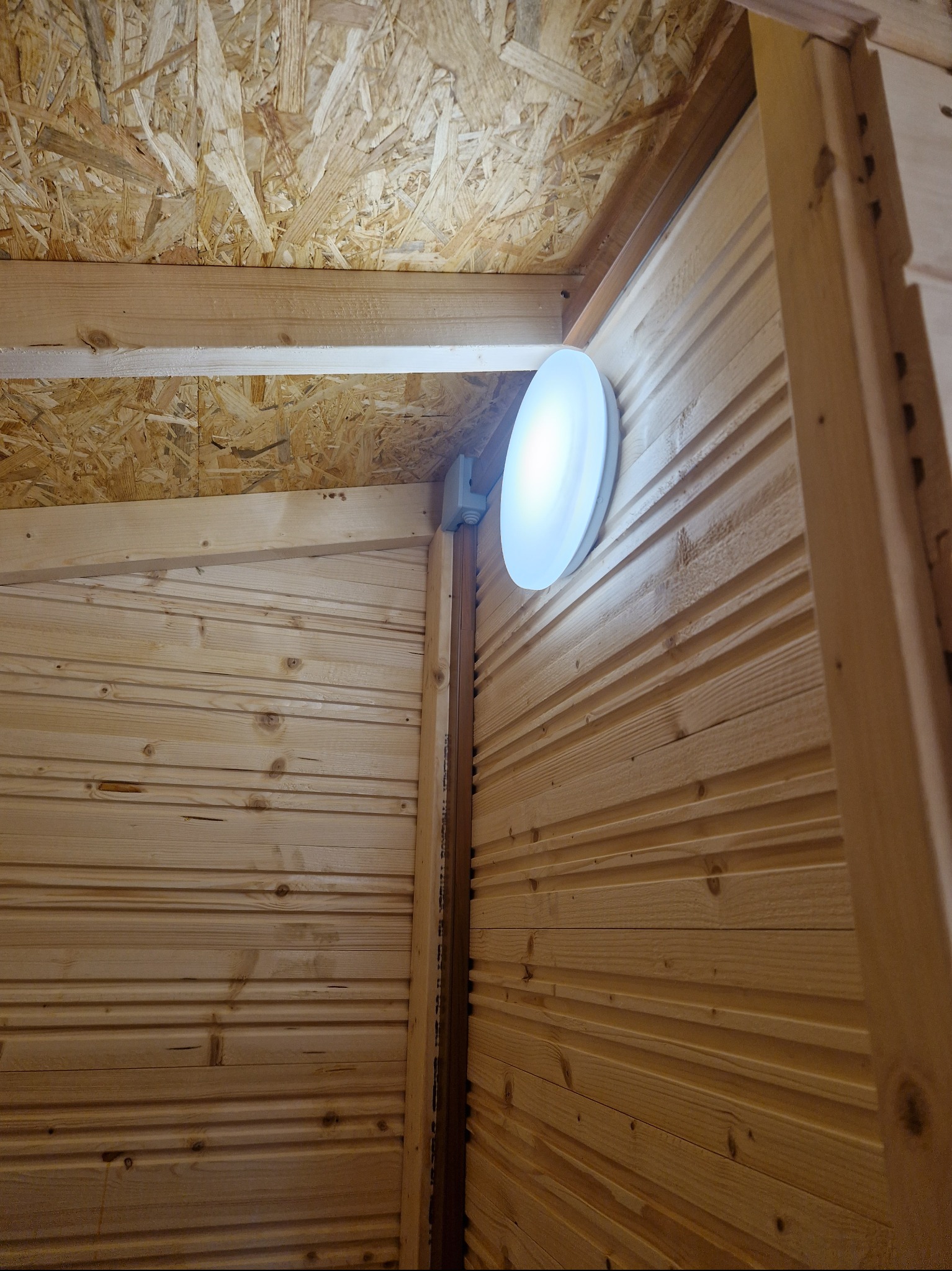 Wnętrze drewnianej konstrukcji z deskami boazeryjnymi i płytą OSB na suficie, oświetlone okrągłą lampą LED zamontowaną na ścianie.