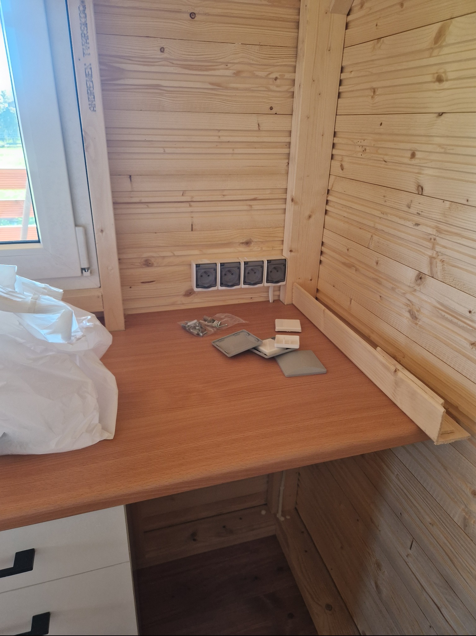 Wnętrze drewnianej konstrukcji w trakcie wykańczania, widoczne gniazdka elektryczne, elementy montażowe i fragment blatu roboczego z szufladami, okno z widokiem na zewnątrz.