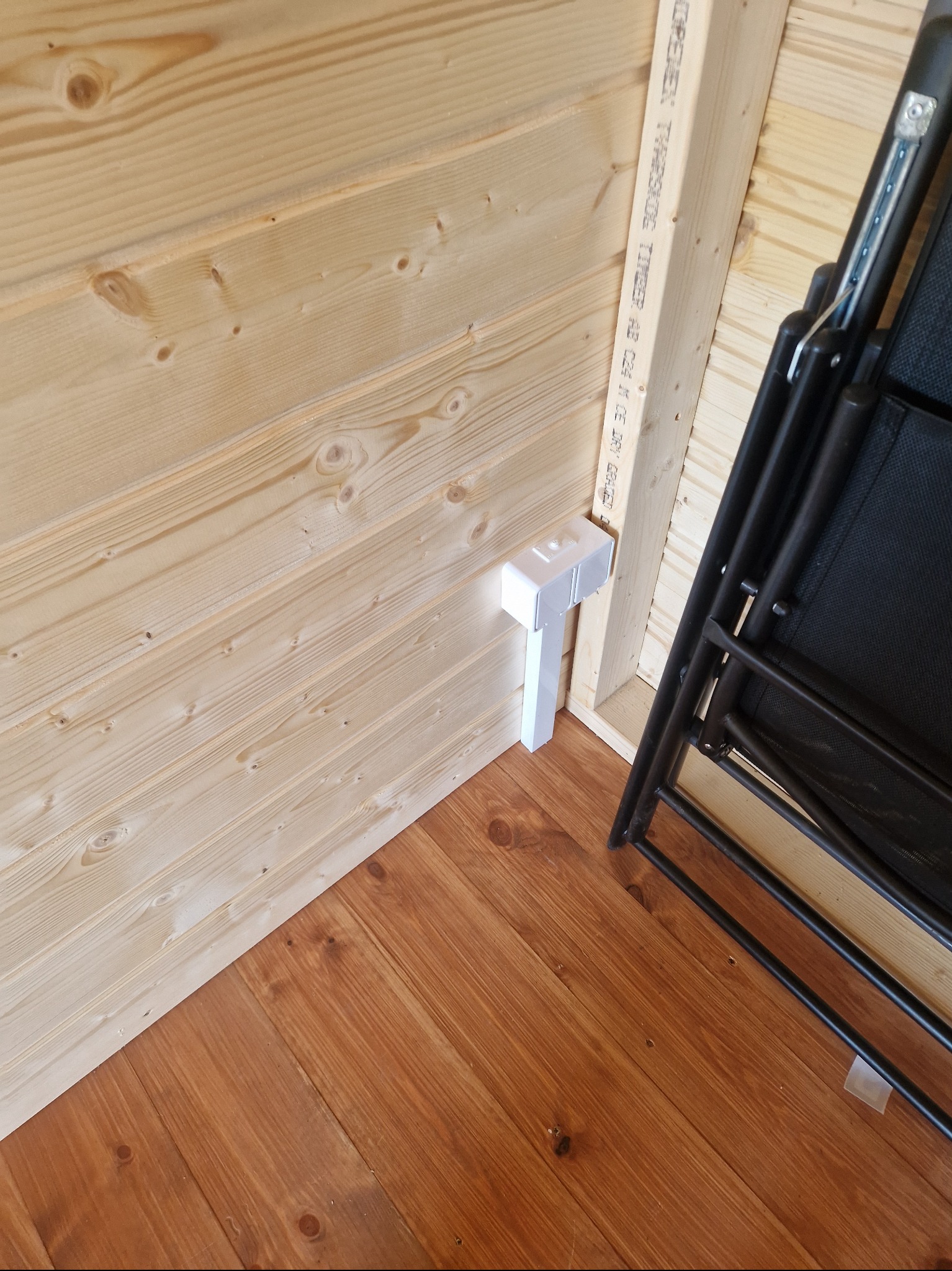 Wnętrze drewnianej konstrukcji z widocznym przyłączem elektrycznym w białej obudowie na ścianie z boazerii oraz fragmentem składanego krzesła.