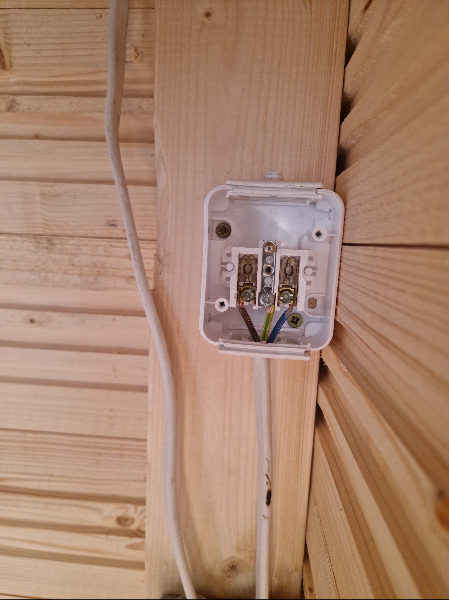 Otwarta, biała puszka elektryczna z widocznymi przewodami wewnątrz, zamontowana na drewnianej ścianie z boazerii.