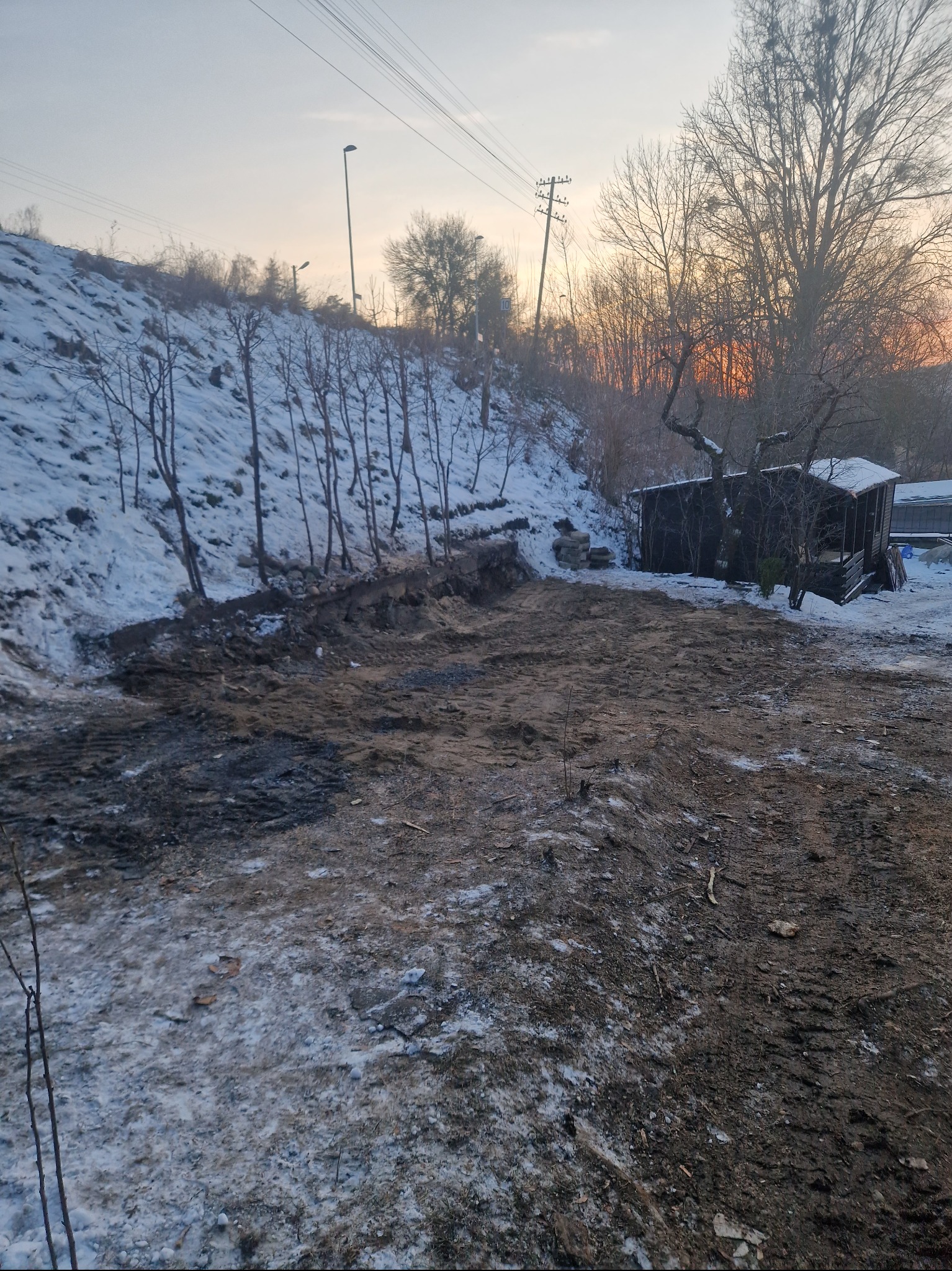Zimowy krajobraz po niwelacji terenu z widoczną ziemią, śniegiem i zarysem zachodzącego słońca na horyzoncie.