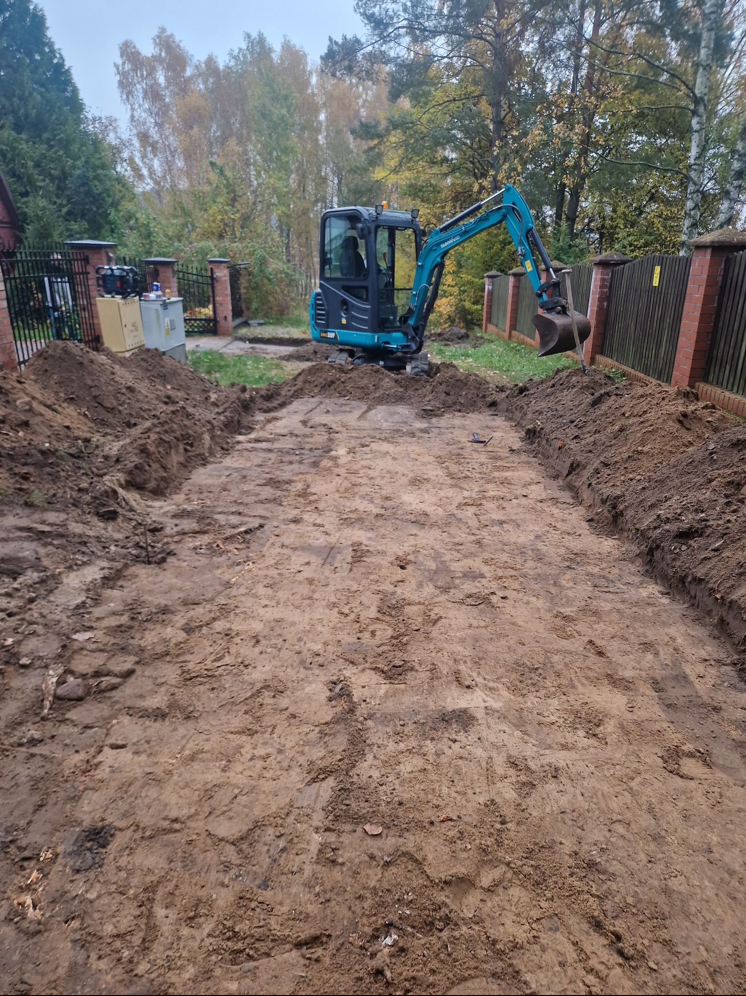 Niebieska mini koparka na działce z wykopanym prostokątnym obszarem ziemi, widoczne ślady gąsienic, w tle ogrodzenie z cegły i drzewa.