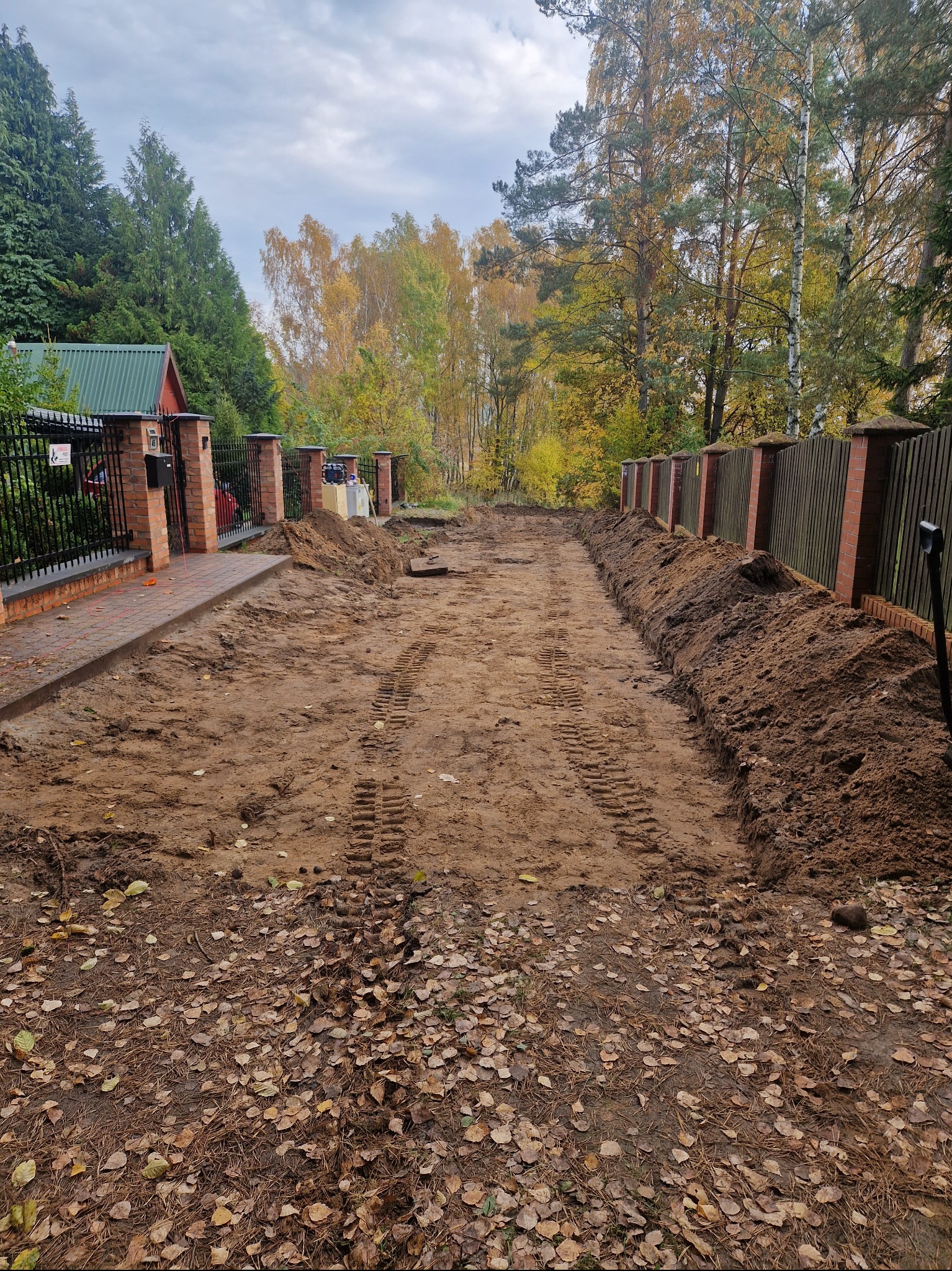Przygotowany teren pod budowę, widoczne ślady opon i wykopane rowy, z brzegami wyznaczonymi przez płot i chodnik z cegły.