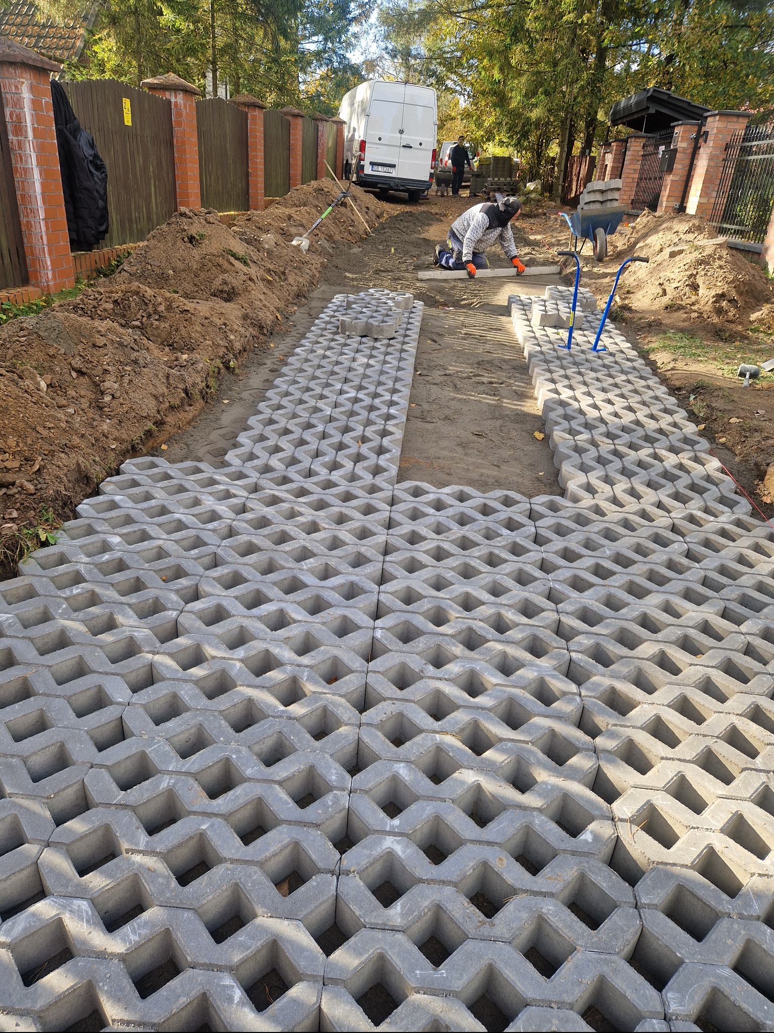 Układanie ażurowych płyt betonowych na przygotowanym podłożu, widoczny pracownik w pomarańczowych rękawicach poziomujący powierzchnię, w tle ziemia, taczka i zaparkowany biały samochód dostawczy.