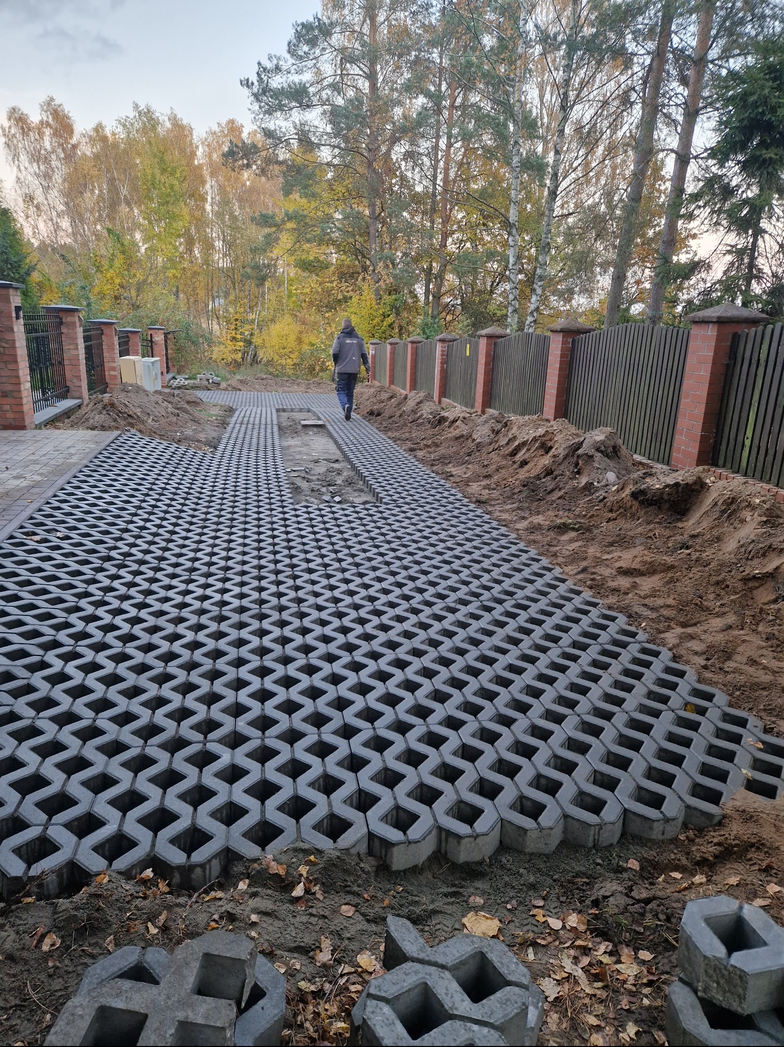 Układanie szarej ażurowej nawierzchni typu kratka na podjeździe w trakcie realizacji, widoczny fragment ogrodzenia z cegły i drewniane sztachety w tle, jesienne drzewa bez liści.